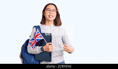 Giovane bella ragazza cinese che indossa zaino studente tenendo Binder e. bandiera britannica sorridente che punta felice con la mano e con il dito Foto Stock