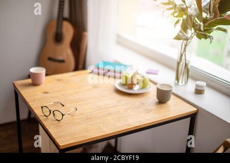 primo piano di un tavolo in legno a casa. scrivania per lavoro freelance. occhiali, tazza di caffè sul tavolo. chitarra sullo sfondo Foto Stock