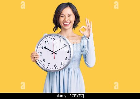 Giovane bella ragazza che tiene grande orologio facendo ok segno con le dita, sorridente amichevole gesturing eccellente simbolo Foto Stock