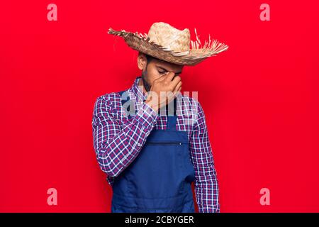 Giovane uomo latino che indossa cappello contadino e grembiule stanco sfregamento naso e occhi sensazione di affaticamento e mal di testa. Stress e frustrazione concetto. Foto Stock