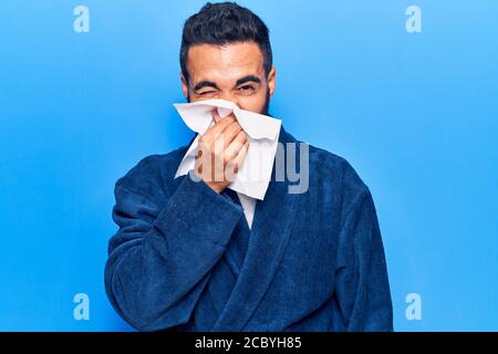Giovane latino uomo malattia usando fazzoletto di carta sul naso. Su sfondo blu isolato Foto Stock