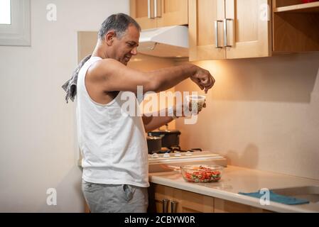 Perfetta vestibilità uomo anziano sorridente e dalla pelle scura in piedi in cucina e preparando l'insalata. Foto Stock