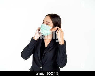 Bella giovane donna asiatica d'affari in vestito nero che indossa una maschera medica verde con la sua mano isolata su sfondo bianco. Foto Stock