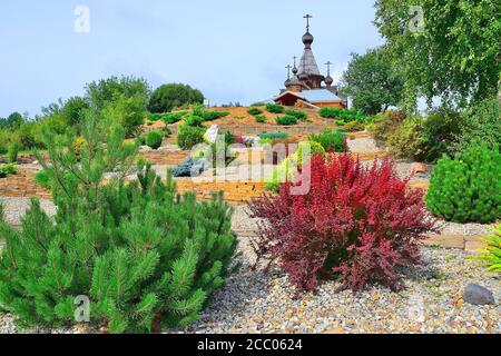 Dwarf alberi, cespugli e fiori sui terrazzi sassosi fortificato da gabions sul pendio collinare - cortile anteriore del tempio ortodosso. Le cupole e croci della chiesa Foto Stock