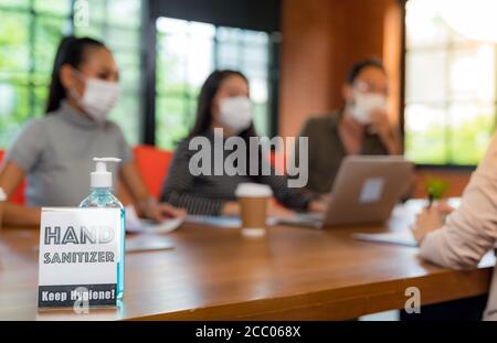 Segnaletica di gel alcolico igienizzante per le mani nella sala riunioni con sfondo di team asiatici che lavorano e si scambiano idee. Indossano una maschera facciale nuova Foto Stock