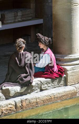 Due donne in costume romano che siedono sul bordo del grande bagno romano, Bath, Inghilterra. Re-enactor femminile con le spalle al pool. Estrema acconciatura. Foto Stock