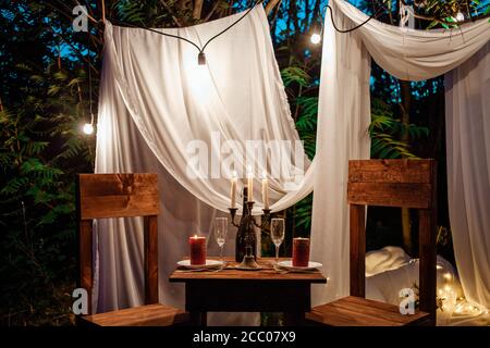 Tavolo nel bosco, cena romantica per due a lume di candela. Tende bianche sull'albero, un ghirlanda di lampadine, bicchieri, vino. Zona foto serale in Foto Stock