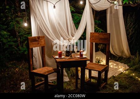 Tavolo nel bosco, cena romantica per due a lume di candela. Tende bianche sull'albero, un ghirlanda di lampadine, bicchieri, vino. Zona foto serale in Foto Stock