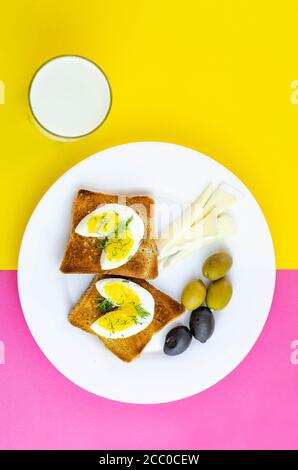 Toast freschi con uova, olive e formaggio di soia per colazione e un bicchiere di latte con su sfondo rosa e giallo. Sano cibo vegetariano concetto Foto Stock