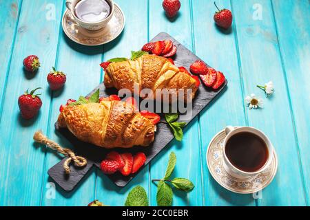 Colazione gustosa. Croissant freschi con fragole e caffè nero su fondo di legno blu Foto Stock