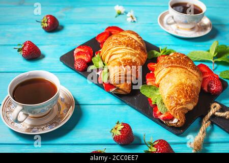 Colazione gustosa. Croissant freschi con fragole e caffè nero su fondo di legno blu Foto Stock