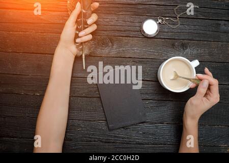 Piuma e tazza di caffè in una mano femminile, notebook e orologio. Vecchio tavolo di legno. Vista dall'alto Foto Stock