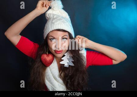 Giovane donna, bruna in un cappello bianco lavorato a maglia, Cute sorride, raddrizza il suo cappello, giocattoli di Capodanno come orecchini, il concetto di congratulazioni per il nuovo anno, foto in studio, sfondo nero. Foto di alta qualità Foto Stock