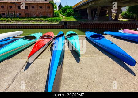 Fila di kayak in plastica rossa e blu a noleggio sono collocati sulla costa di cemento accanto al fiume. Foto Stock