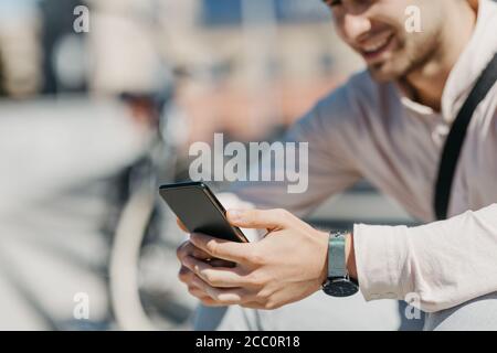 Online in città. Sorridente ragazzo in camicia si siede in strada e guarda lo smartphone Foto Stock