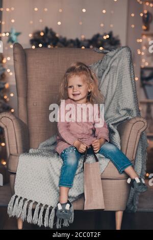 Carino divertente bambina di 3-4 anni indossando pantaloni in denim e maglia maglione seduta in sedia sopra Natale luci incandescenti in guardaroba camera. Vacanze invernali Foto Stock