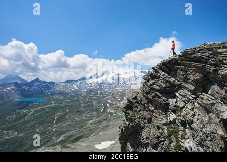 Viaggiatore maschile in piedi sul bordo di alta scogliera rocciosa sotto il bel cielo nuvoloso. Uomo alpinista ammirando la vista della valle di montagna con colline erbose e lago blu. Concetto di viaggio e trekking. Foto Stock