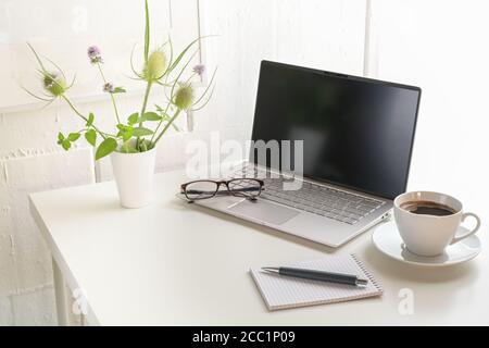 Scrivania da ufficio bianca con computer portatile, bicchieri, blocco note, bouquet di fiori e una tazza di caffè, piccolo luogo di lavoro a casa o in una moderna azienda contro una parete luminosa Foto Stock