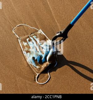 closeup di qualcuno che utilizza un estensione reach per raccogliere un maschera chirurgica blu utilizzata gettata sulla sabbia bagnata di il mare o riportato dall'oceano a th Foto Stock