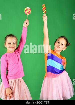 I bambini mangiano grandi caramelle dolci colorate. Ragazze con facce felici mettono lollipop su sfondo verde. Sorelle con lollipop rotondi e lunghi. Concetto di pasticceria e infanzia. Foto Stock