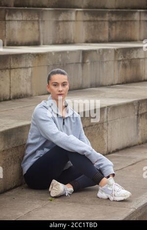 Una giovane donna sportiva che indossa abbigliamento sportivo seduto davanti a una serie di gradini, pronta per l'esercizio. Foto Stock