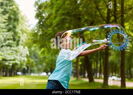 Vita ritratto di carino afroamericano ragazzo giocando con bolle all'aperto nel parco verde tenendo grande bacchetta bolla, copia spazio Foto Stock