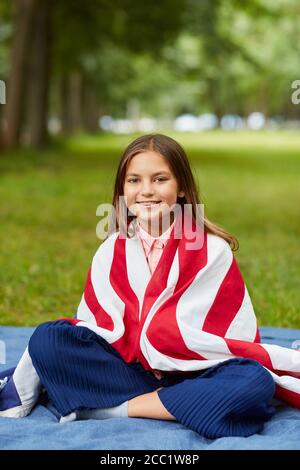 Verticale ritratto a lunghezza intera di carina ragazza coperta da bandiera americana seduta su coperta da picnic nel parco e sorridente a macchina fotografica, copia spazio Foto Stock