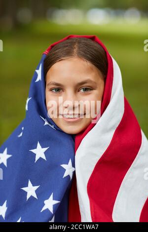 Verticale pieno primo piano su ritratto di cute ragazza coperto da Bandiera americana che sorride alla macchina fotografica mentre si siede su erba verde all'aperto Foto Stock