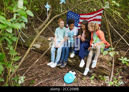 Gruppo multietnico di bambini che usano il tablet digitale mentre giocano nel cortile nascosto sotto i cespugli con la bandiera americana sullo sfondo, spazio di copia Foto Stock