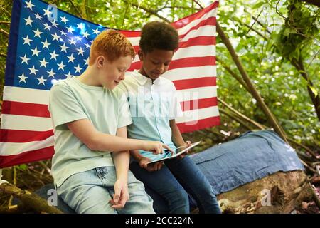 Ritratto di due ragazzi adolescenti che usano il tablet digitale all'aperto mentre nascondendosi sotto i rami del grande albero nella foresta o giocando nel cortile posteriore Foto Stock