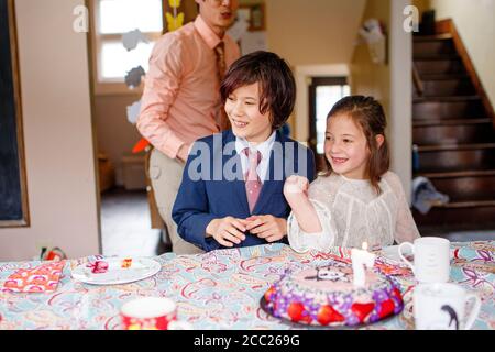 I fratelli sorridenti si siedono a tavola in abiti eleganti con illuminati torta di compleanno Foto Stock