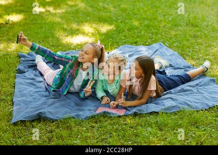 Ritratto ad alto angolo di tre ragazze adolescenti che prendono selfie insieme mentre giacciono su erba verde in un parco all'aperto illuminato dalla luce del sole, copia spazio Foto Stock