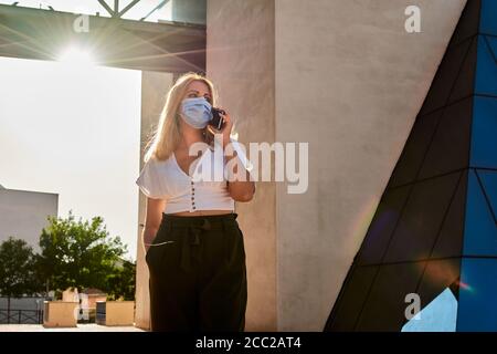 Giovane donna parla al telefono mentre indossa una maschera Foto Stock