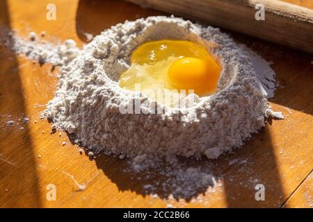 Ingredienti per la preparazione dell'impasto. Farina setacciata e uovo spezzato su tavola di legno e coppiglia. Foto di alta qualità Foto Stock