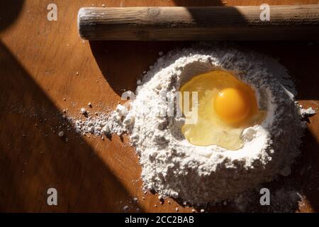 Ingredienti per la preparazione dell'impasto. Farina setacciata e uovo spezzato su tavola di legno e coppiglia. Foto di alta qualità Foto Stock