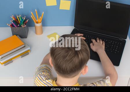 Ragazzo che usa il suo laptop per studiare online durante la quarantena, seduto al tavolo contro la parete blu. Homeschooling, corsi di formazione online o formazione Foto Stock