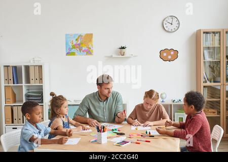 Vista ad alto angolo di un insegnante di sesso maschile che lavora con un gruppo multietnico di bambini che disegnano immagini durante la lezione d'arte a scuola o al centro di sviluppo, copy sp Foto Stock