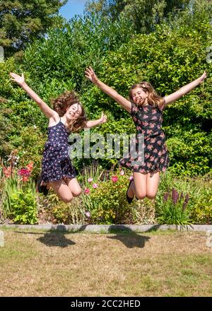 Ritratto verticale di due giovani ragazze che saltano con eccitazione con le loro braccia distese in giardino. Foto Stock