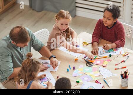 Vista ad alto angolo su un gruppo multietnico di bambini che disegnano immagini con pastelli mentre si godi l'arte e la classe artigianale in pre-scuola o centro di sviluppo, co Foto Stock