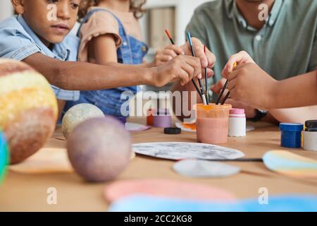 Primo piano di un gruppo multietnico di bambini che tengono pennelli e dipingono il modello di pianeta mentre si tengono lezioni di arte e artigianato a scuola o in un centro di sviluppo, Foto Stock
