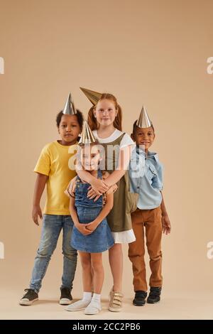 Ritratto verticale a tutta lunghezza di diversi gruppi di bambini che indossano cappelli da festa che guardano la fotocamera mentre si levano in piedi su uno sfondo normale in studio Foto Stock