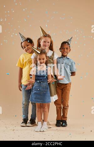 Ritratto verticale a tutta lunghezza di diversi gruppi di bambini che indossano i cappelli da festa sorridono davanti alla fotocamera mentre si levano in piedi sotto i colorati confetti scoppiare in studio Foto Stock