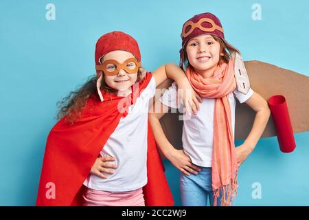 Due giocosi bambini caucasici in tuta pilota, mantello e aeroplano di cartone si trovano isolati su sfondo blu Foto Stock