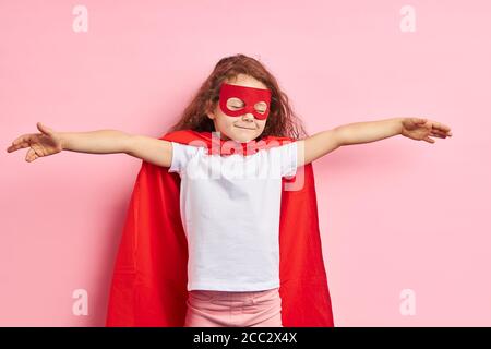 Bella bambina brillare fuori indossando vestito rosso supereroe e maschera rossa, isolato sfondo rosa Foto Stock