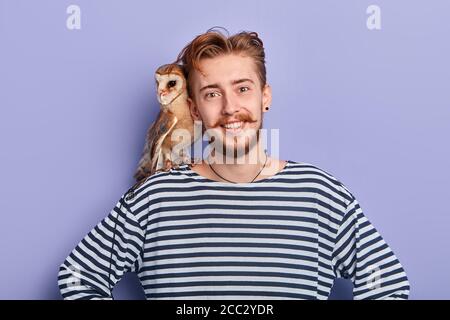 sorridente ragazzo positivo con le mani sui fianchi offre di scattare una foto con il suo uccello, business, vendita concetto. isolato sfondo blu, studio shot Foto Stock