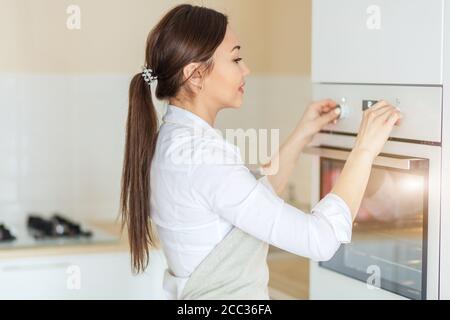 Allegra ragazza asiatica che accende il forno in cucina. Primo piano vista laterale foto. Chef ha messo il piatto nel forno per preriscaldarlo Foto Stock