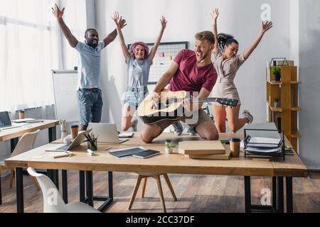 Fuoco selettivo delle persone di affari multietniche che saltano mentre i colleghi giocano chitarra acustica in ufficio Foto Stock
