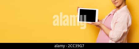 vista ritagliata di una giovane donna incinta che tiene un tablet digitale con schermo vuoto su un'immagine gialla orizzontale Foto Stock