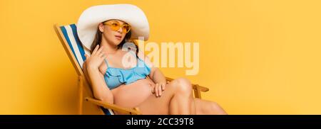 foto panoramica di donna incinta sognante in costume da bagno e paglia cappello che prende il sole in sedia a sdraio sul giallo Foto Stock