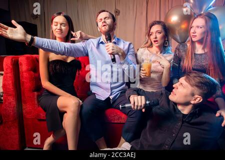 i giovani e felici amici caucasici trascorrono il tempo di festa nel karaoke bar, cantano e si divertono insieme, indossando abiti da festa Foto Stock